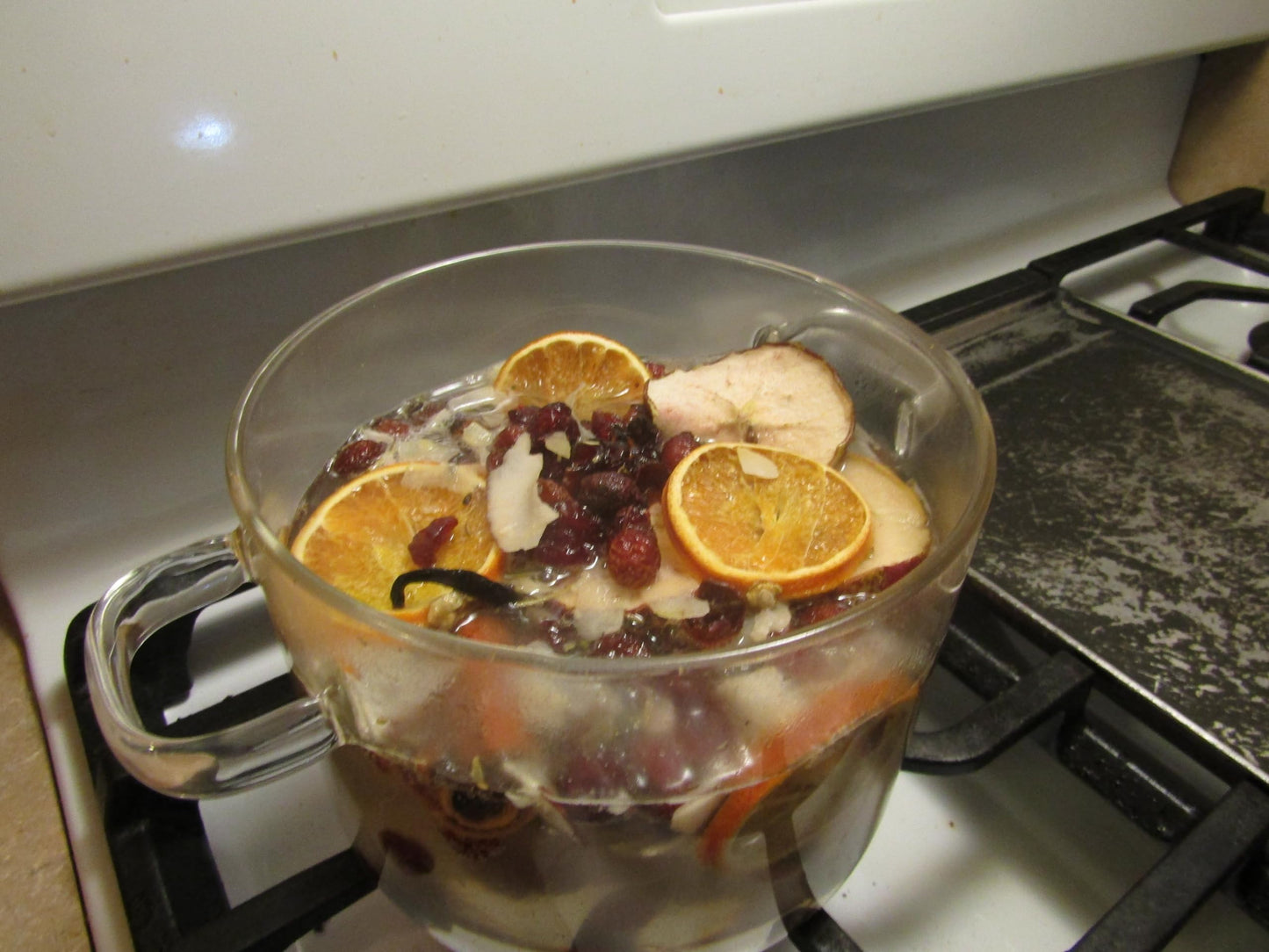 Glass pot on stove with simmering potpourri with oil including dried orange slices and cranberries