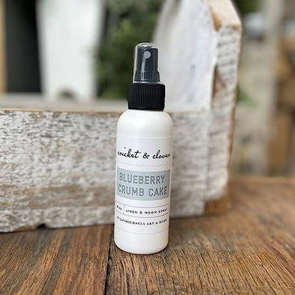Cricket and Clover Blueberry Crumb Cake Potpourri Room Spray white bottle with black spray top on rustic wooden surface