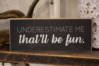 Black wooden home decor sign with white text underestimate me that will be fun sign on rustic shelf